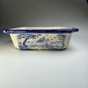 May include: Rectangular ceramic bonsai pot with a cream-colored base and a blue rim. The pot features a blue and white landscape design with trees, mountains, and a bridge. The pot has a rectangular shape with a slightly flared rim.