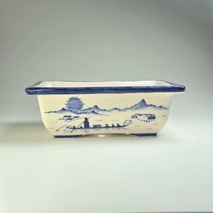 May include: Cream-coloured rectangular ceramic planter with blue trim. The planter is decorated with a hand-painted landscape scene featuring mountains, a boat, and buildings, all in shades of blue.