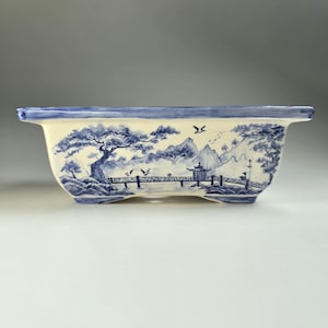 May include: A rectangular ceramic planter with a blue and white landscape design. The planter features a bridge, trees, mountains, and birds. The rim is painted blue. The planter is elevated on small feet.