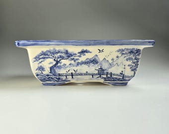 May include: A rectangular ceramic planter with a blue and white landscape design. The planter features a bridge, trees, mountains, and birds. The rim is painted blue. The planter is elevated on small feet.