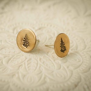 May include: A pair of gold-colored stud earrings. Each earring features a small, oval-shaped design with a detailed black tree silhouette in the center. The earrings are set against a white, textured background.