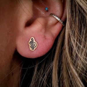 Op de afbeelding: Close-up van een oor met een goudkleurige slangenoorbel. De oorbel heeft een zwarte omtrek en een gedetailleerd slangenontwerp. Een zilveren oorring en een kleine turkooizen stud zijn ook zichtbaar.