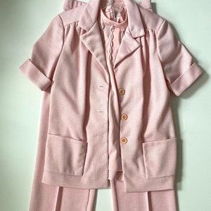 May include: A light pink vintage suit set, including a short-sleeved blazer with large pockets and a matching pair of pants. The blazer has three buttons and a collared neckline. A light pink blouse with a decorative neckline is visible underneath.