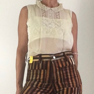 Puede incluir: Una blusa sin mangas de color crema con un panel frontal y cuello con detalles de encaje. La blusa se combina con pantalones a rayas verticales marrones, bronceados y dorados. Los pantalones tienen cierre de botón y trabillas para el cinturón.