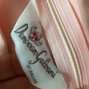 May include: Close-up of a pink garment with a zipper and a white label. The label reads "Donovan Galvani of Dallas" in cursive script. The zipper is light pink with a beige center.