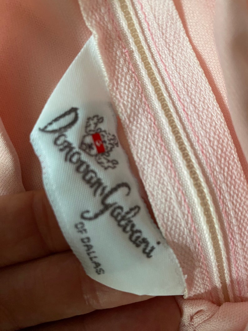May include: Close-up of a pink garment with a zipper and a white label. The label reads "Donovan Galvani of Dallas" in cursive script. The zipper is light pink with a beige center.