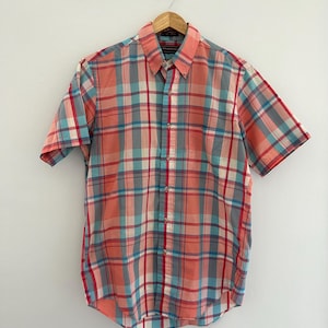 May include: A short-sleeved button-down shirt with a plaid pattern in coral, gray, red, and blue. The shirt is hanging on a wooden hanger against a white background. The shirt has a button-down collar.