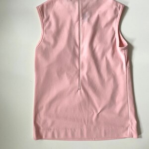 May include: A sleeveless, pale pink top with a zipper down the back. The top has a simple, minimalist design with a round neckline and a straight hemline. The fabric appears smooth and has a slight sheen.