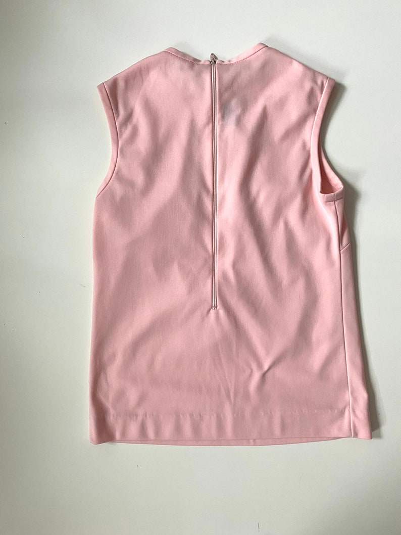 May include: A sleeveless, pale pink top with a zipper down the back. The top has a simple, minimalist design with a round neckline and a straight hemline. The fabric appears smooth and has a slight sheen.