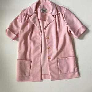 May include: A light pink blazer with short sleeves, two front pockets, and three buttons. The blazer has a collared neckline and a label at the neck. The fabric has a subtle textured appearance.