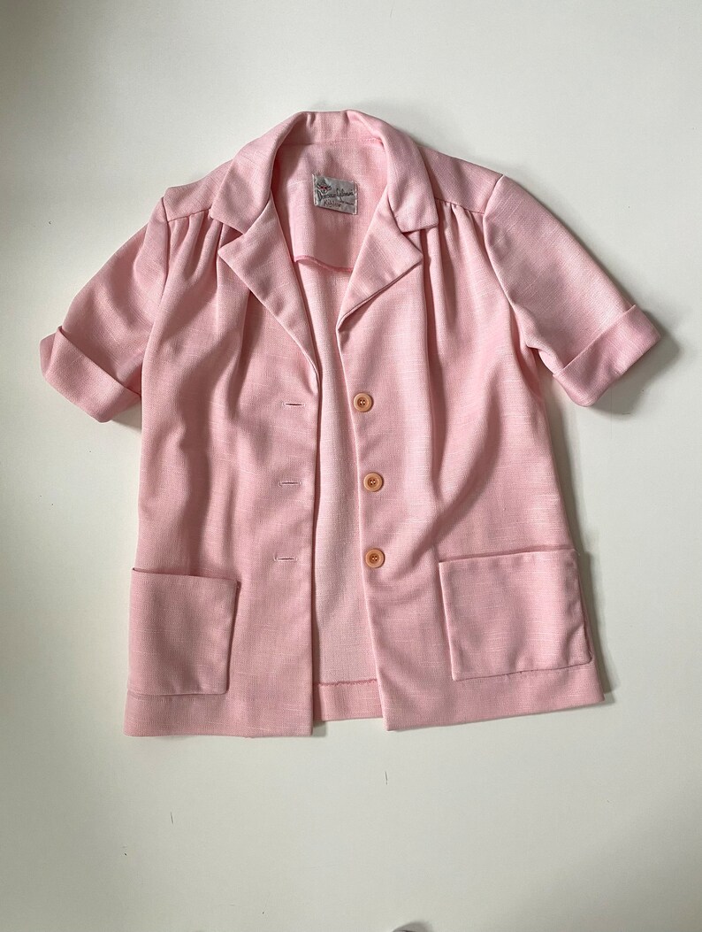 May include: A light pink blazer with short sleeves, two front pockets, and three buttons. The blazer has a collared neckline and a label at the neck. The fabric has a subtle textured appearance.