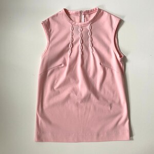 May include: A sleeveless, light pink top with a round neckline and a vertical zipper. The front features a series of decorative, stitched oval shapes. The top has a simple, tailored design, suitable for various outfits.