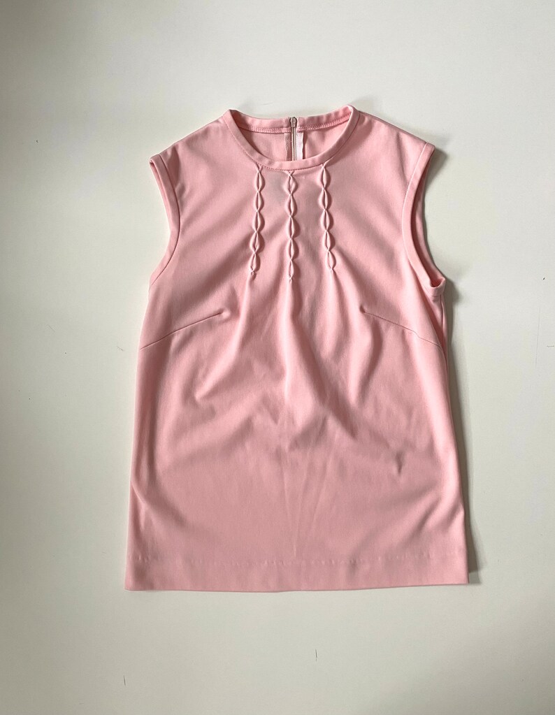 May include: A sleeveless, light pink top with a round neckline and a vertical zipper. The front features a series of decorative, stitched oval shapes. The top has a simple, tailored design, suitable for various outfits.