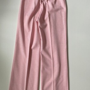 May include: Pale pink wide-leg pants. The pants have a high waist and a subtle textured appearance. The fabric appears to be a lightweight material, suitable for warmer weather. The pants are neatly pressed with a center crease down each leg.