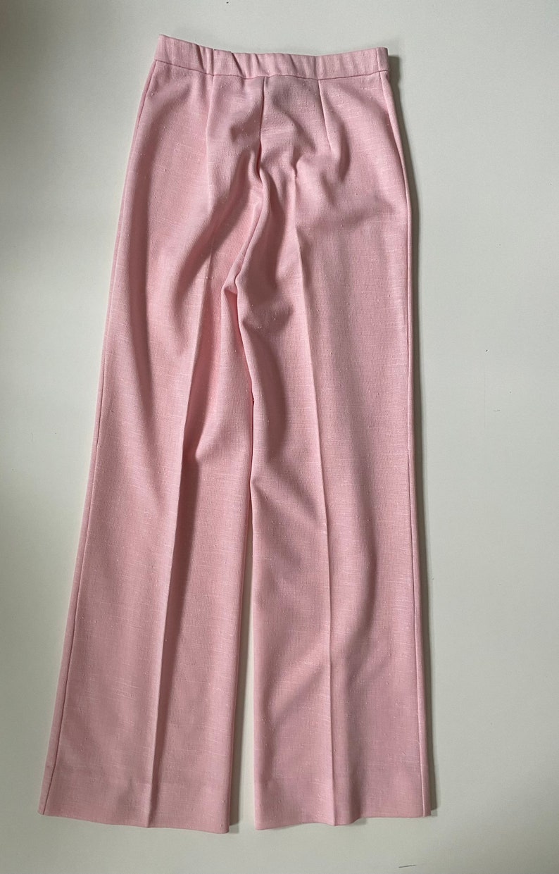 May include: Pale pink wide-leg pants. The pants have a high waist and a subtle textured appearance. The fabric appears to be a lightweight material, suitable for warmer weather. The pants are neatly pressed with a center crease down each leg.