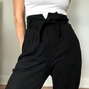 May include: Black high-waisted pants with a matching fabric belt tied in a bow. The pants have a wide waistband and a relaxed fit. The model is wearing a white tank top. The pants are made of a soft, flowing fabric.