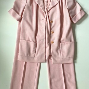 May include: A light pink three-piece suit set. The set includes a short-sleeved blazer with three buttons and two front pockets, a matching pair of wide-leg pants, and a collared blouse. The suit is made of a textured fabric.