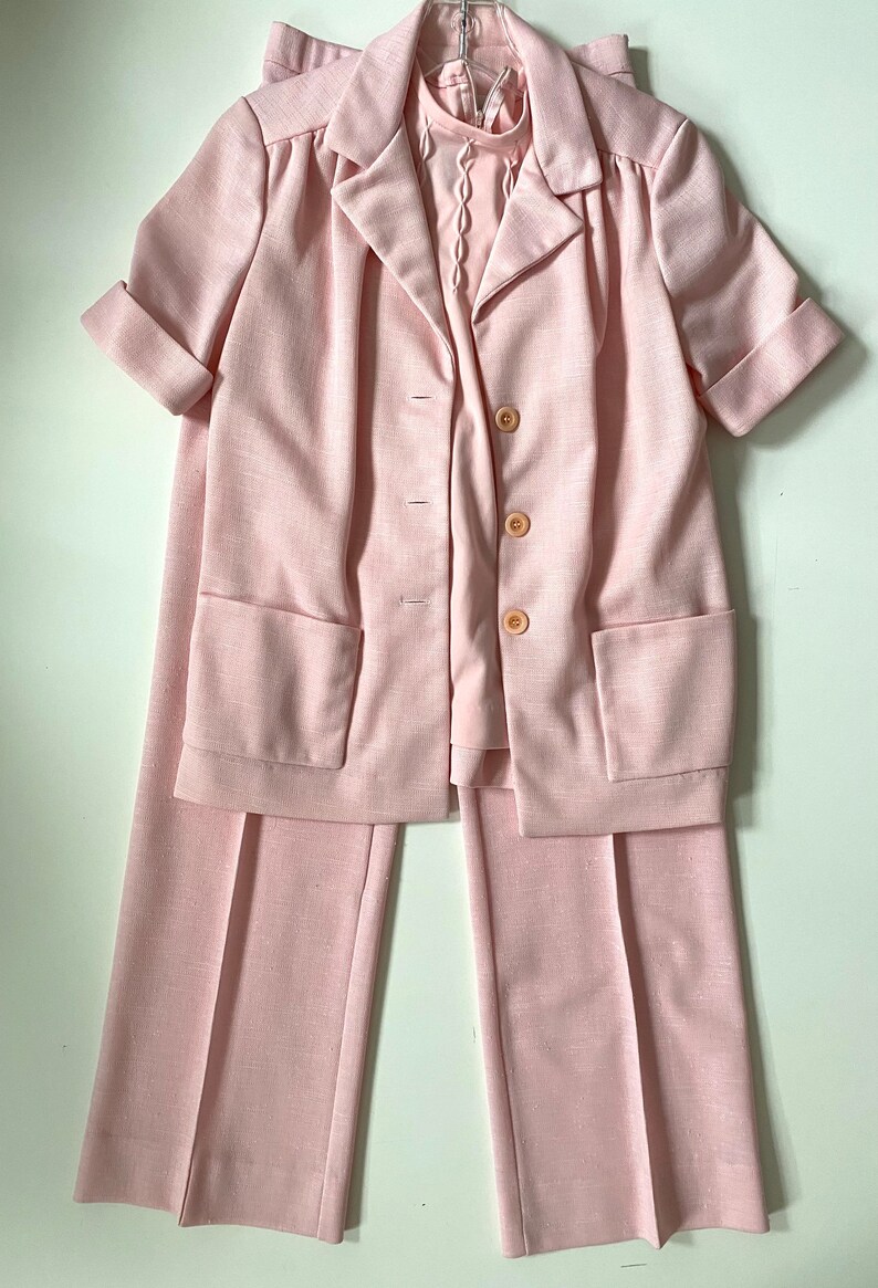 May include: A light pink three-piece suit set. The set includes a short-sleeved blazer with three buttons and two front pockets, a matching pair of wide-leg pants, and a collared blouse. The suit is made of a textured fabric.