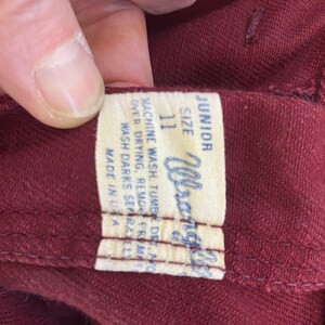 Może przedstawiać: Zbliżenie bordowego ubrania z białą metką. Na metce widnieje napis "JUNIOR SIZE 11 Wrangler" oraz instrukcje prania. Ubranie jest wyprodukowane w USA.