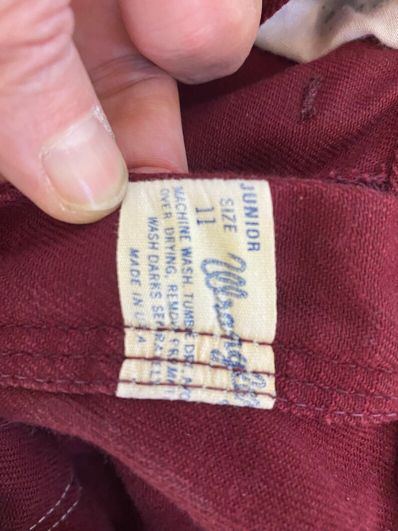 Może przedstawiać: Zbliżenie bordowego ubrania z białą metką. Na metce widnieje napis "JUNIOR SIZE 11 Wrangler" oraz instrukcje prania. Ubranie jest wyprodukowane w USA.