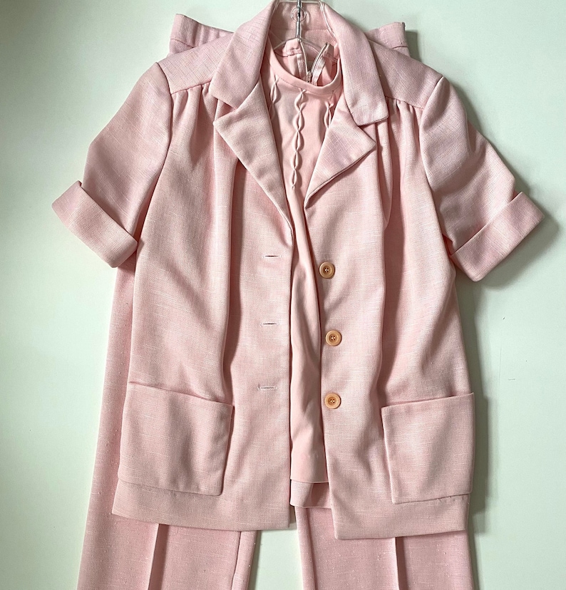 May include: A light pink vintage suit set, including a short-sleeved blazer with large pockets and a matching pair of pants. The blazer has three buttons and a collared neckline. A light pink blouse with a decorative neckline is visible underneath.