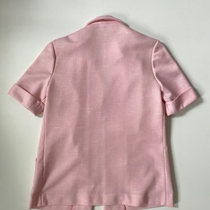 May include: A light pink short-sleeved blazer with a notched collar and folded cuffs. The blazer has a textured fabric and a split hem at the back. The garment is displayed on a white surface.