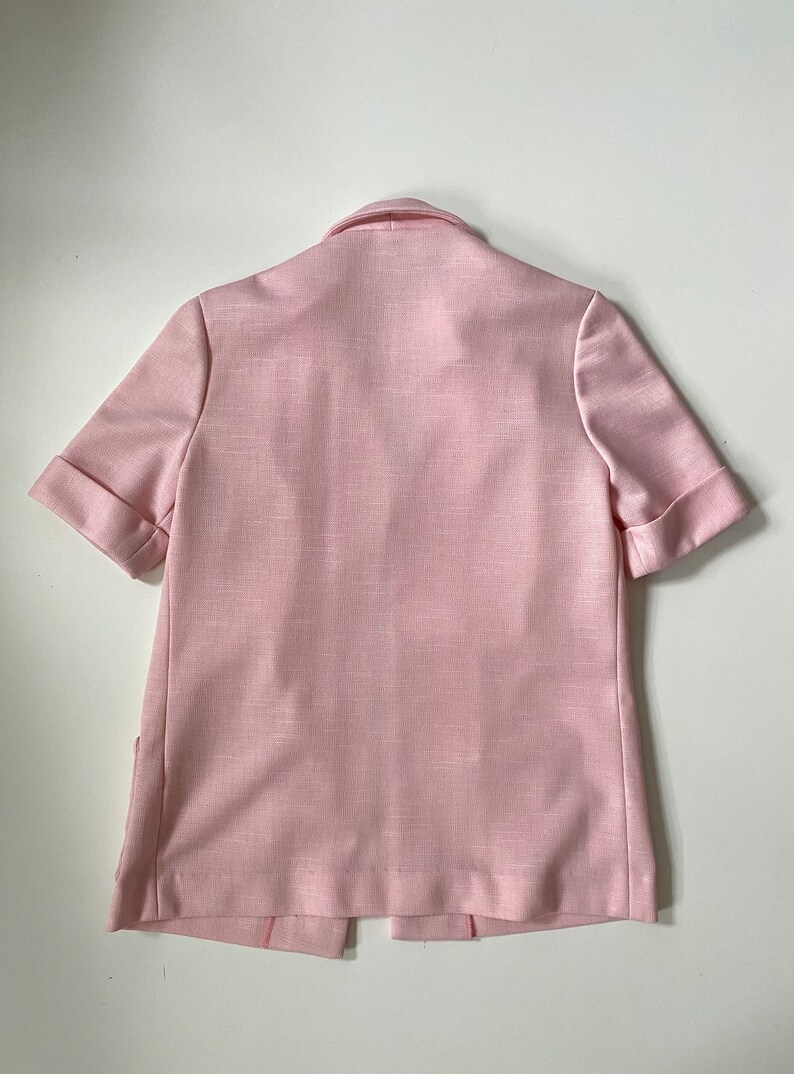 May include: A light pink short-sleeved blazer with a notched collar and folded cuffs. The blazer has a textured fabric and a split hem at the back. The garment is displayed on a white surface.