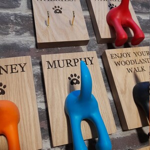 May include: Wooden wall hooks shaped like dog tails in various colours, including blue, orange, and red. The hooks are labelled with names like "Murphy", "Ney", and "Max". One hook has the text "Let's go walkies!" and another has the text "Enjoy your woodland walk".