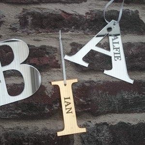 May include: Three silver mirror-like ornaments shaped as the letters A, B, and I. The letter A has the name "Alfie" engraved on it, the letter B has the name "Ben" engraved on it, and the letter I has the name "Ian" engraved on it. The ornaments are hanging from white ribbons against a brick wall background.