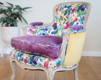 May include: A vintage armchair with a floral patterned fabric upholstery. The chair has a light gray painted finish and features a purple velvet cushion. The chair is sitting on a wooden floor.