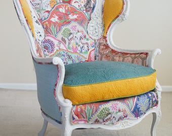 May include: A white ornate wingback chair with a colorful patterned fabric back and seat. The seat cushion is teal with a yellow border. The chair's arms are yellow and blue. The fabric features a whimsical design with animals and floral elements.