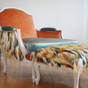May include: A white, ornate armchair with a tufted orange velvet seat and a teal velvet cushion. The chair has a fluffy, multi-colored fringe trim. A matching footstool with a fluffy, multi-colored top sits in front of the chair.