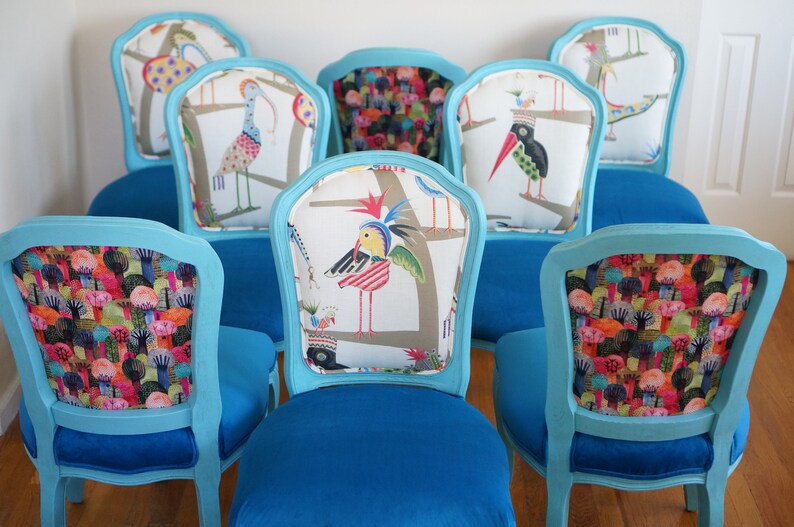 May include: Six turquoise blue dining chairs with different colorful patterned fabric backrests. The chairs have a classic design with a curved back and a padded seat.