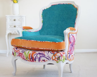 May include: A white-framed armchair with a teal back and seat, and orange trim. The seat cushion is orange. The chair's base and sides feature a colorful floral pattern. A small white side table and vase of flowers are in the background.