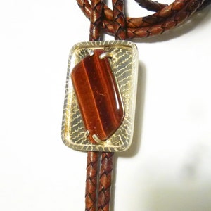 May include: A brown leather bolo tie with a silver rectangular pendant featuring a red tiger's eye stone.