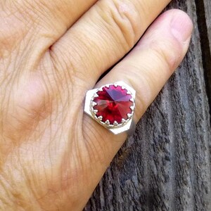 May include: A silver ring with a large, faceted, deep red gemstone. The gemstone is set in a geometric, silver bezel. The ring is on a person's finger, with a wooden background.