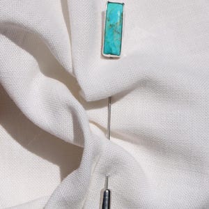 May include: A silver-toned lapel pin with a rectangular turquoise stone. The pin is set against a white fabric background, highlighting the vibrant blue-green color of the stone. The pin is a stylish accessory.