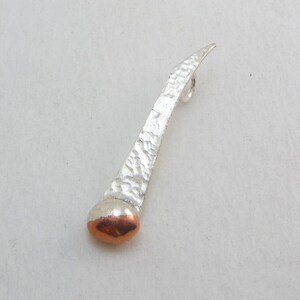 May include: A silver pendant with a hammered texture and a copper ball at the bottom. The pendant has a loop at the top for attaching a chain.