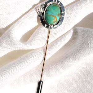May include: A silver-toned stick pin with an oval turquoise stone and a small, clear accent stone. The pin has a gold-colored shaft and a black, rectangular tip. The pin is set against a white, textured background.