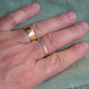 May include: A hand wearing two gold rings. The ring on the index finger is a simple band, while the ring on the middle finger is a wider band with a textured design.