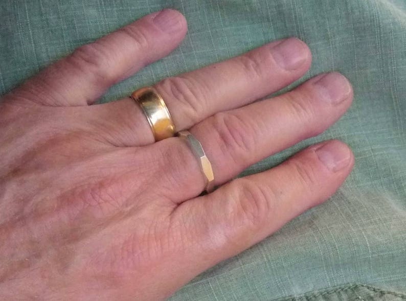 May include: A hand wearing two gold rings. The ring on the index finger is a simple band, while the ring on the middle finger is a wider band with a textured design.