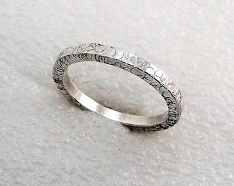 May include: A silver band ring with a textured surface. The ring is simple and elegant.