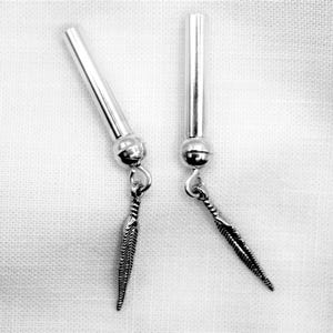 May include: A pair of silver-toned earrings. Each earring features a long, cylindrical post connected to a small ball and a feather-shaped charm. The earrings are displayed against a white, textured background.