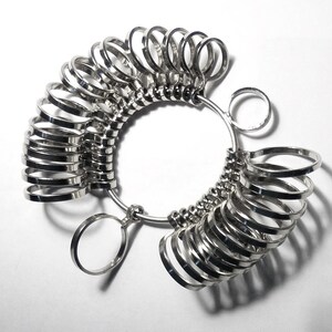 May include: A set of silver ring sizers arranged in a circular pattern. Each ring is a different size, attached to a metal loop. The sizers are on a white background, with shadows adding depth. This tool is used for measuring finger sizes.