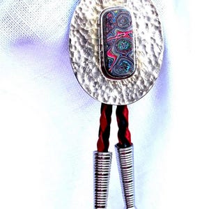 May include: Silver bolo tie with an oval, hammered-texture pendant. The pendant features a rectangular, colorful stone with swirls of red, blue, and green. The tie has a black and red braided cord and silver tips.