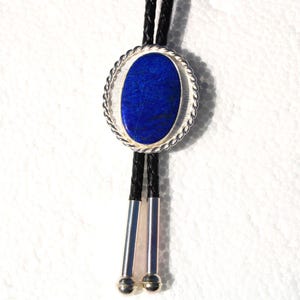 May include: A bolo tie featuring a large, oval, deep blue stone set in a silver-toned, rope-edged frame. The tie has a black braided cord with silver-toned metal tips and a decorative slide. The background is white.