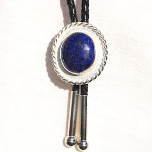 May include: A bolo tie featuring a dark blue oval stone set in a silver-toned frame with a twisted rope design. The tie has a black braided cord and silver-colored tips.