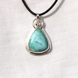 May include: A silver pendant with a triangular, light blue and green larimar stone. The stone is set in a silver bezel and hangs from a black cord. A small star-shaped detail adorns the top of the pendant.