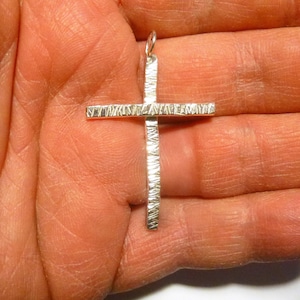 May include: A silver cross pendant with a textured surface. The cross is simple and elegant, with a slightly rounded shape.