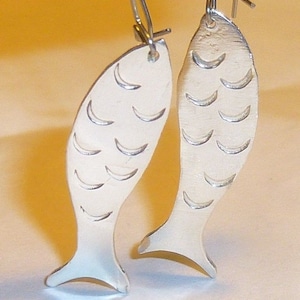 May include: A pair of silver fish-shaped earrings with a textured surface. The earrings are hanging from silver hooks.